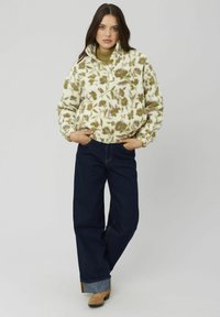 Pull en peluche crème avec motif floral vert et violet, présentant un col à demi-zip, associé à un jean large bleu foncé.