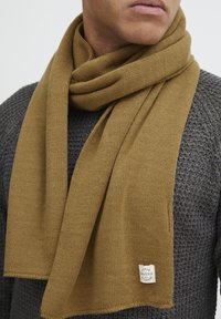 Strikket tørklæde i en varm brun farve, lavet af blødt materiale, har en subtil tekstur og et lille mærke med detaljen "Denim Blend".