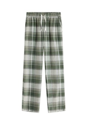 Junge steht barfuß, trägt ein weißes oversized T-Shirt und grün-grau karierte Hose, die Hände in den Taschen, vor schlicht weißem Hintergrund.