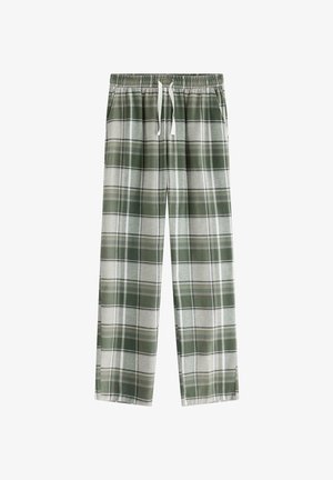 Garçon debout pieds nus portant un t-shirt blanc oversize et un pantalon à carreaux verts et gris, les mains dans les poches, sur fond blanc uni.