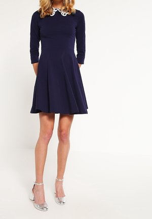 Femme portant une robe bleu marine longueur genou avec manches trois-quarts et col en dentelle blanche, associée à des escarpins argentés à bride autour de la cheville.