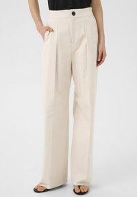 Beige wide-leg trousers in a textured fabric, featuring a front button closure, pleats, and side pockets, styled with a black top.