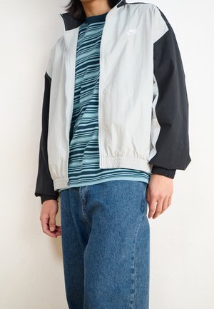 Person iført en sort og hvid Nike-jakke over en grønstribet skjorte og løse blå jeans, stående mod en ensfarvet lys baggrund.