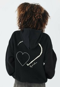 Felpa nera con un grande cuore bianco stampato e un design a linea curva sul retro. Presenta un cappuccio con coulisse e un materiale morbido e testurizzato.