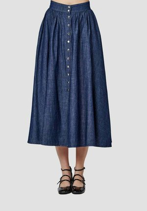 Jupe midi en jean bleu foncé taille haute avec ouverture boutonnée sur le devant, portée avec des chaussures noires en cuir verni à brides sur un fond neutre.