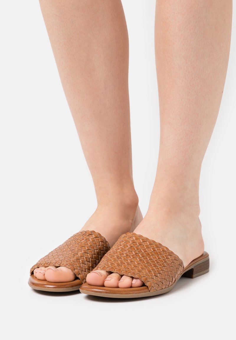 Pieds portant des sandales marron en cuir tressé, à enfiler, avec de petits talons, sur fond blanc.