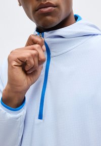 Άτομο που φοράει λευκό μπουφάν με μικρές οπές, κλείνοντας το φωτεινό μπλε φερμουάρ στο μπροστινό μέρος κοντά στον λαιμό και την κουκούλα.