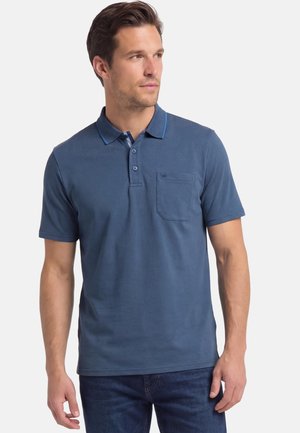 Man in een blauw poloshirt met korte mouwen, knopen en een borstzak, gecombineerd met een blauwe spijkerbroek, kijkt opzij.