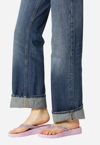 Pies con chanclas moradas texturizadas combinadas con jeans de denim azul descoloridos y remangados sobre un fondo liso.
