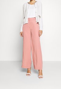 Biały, puszysty kardigan z guzikami, w połączeniu z jasnoróżowymi spodniami o szerokich nogawkach. Modelka nosi białe sandały na obcasie. Prosty i nowoczesny design.