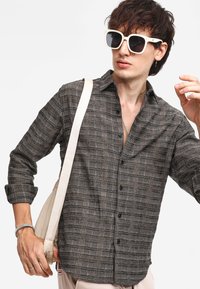 Camicia grigia a scacchi realizzata in tessuto intrecciato, con bottone anteriore e maniche arrotolate. Gli accessori includono una borsa leggera e occhiali da sole oversize.