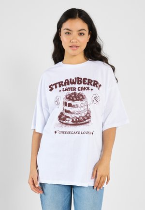 T-shirt blanc avec un design graphique d'un gâteau aux fraises, accompagné de texte. Manches courtes, coupe oversize, en coton décontracté.