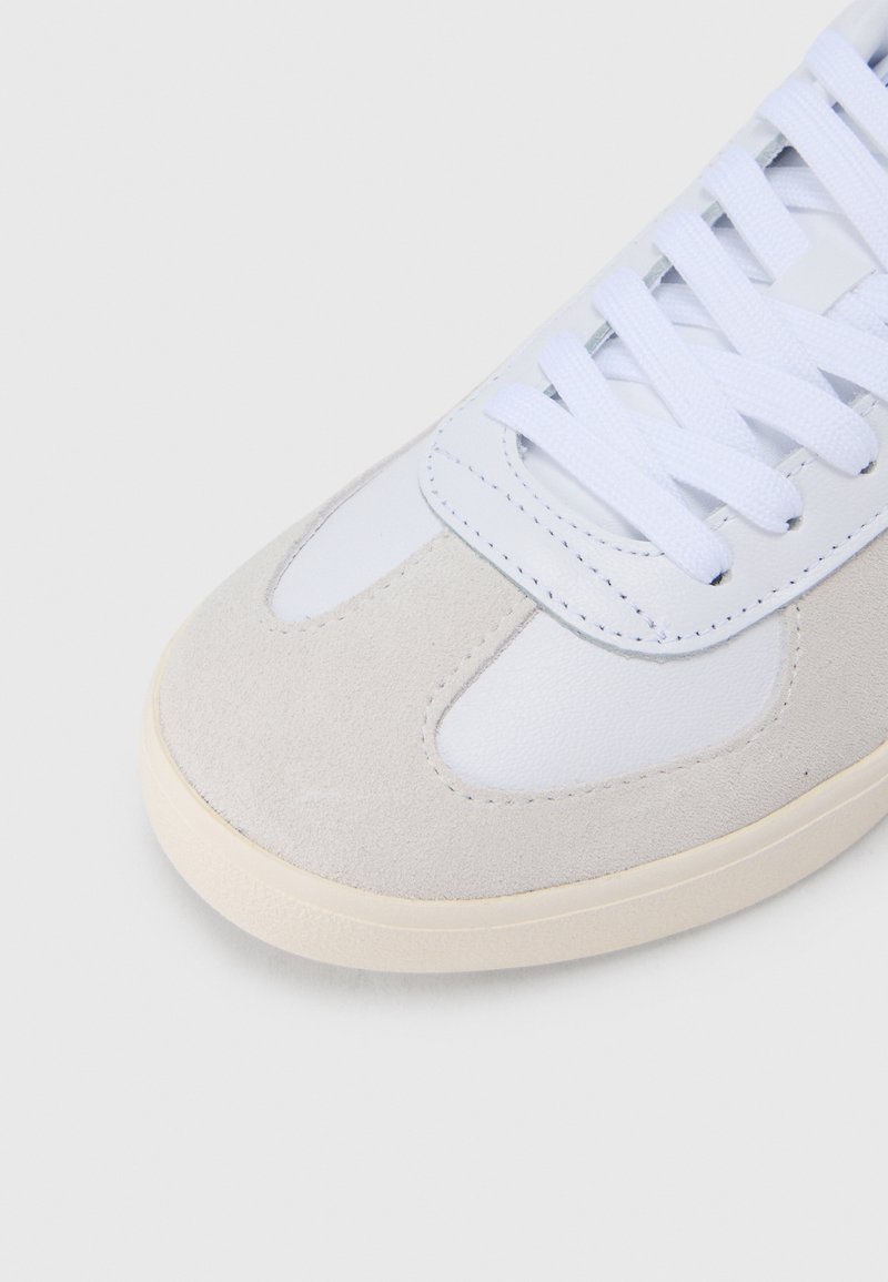 Nærbillede af en beige ruskinds- og hvid lædersneakersnude med hvide snørebånd på en lys baggrund.