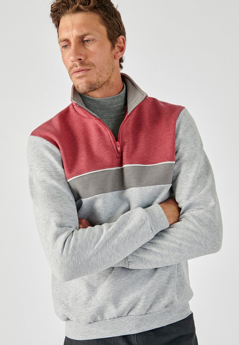 Sudadera gris con cremallera que presenta una franja horizontal roja y gris, de textura suave, cuello alto y puños acanalados. Diseño casual con bloque de color.