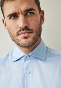 Chemise de couleur bleu clair avec un col évasé, deux boutons blancs et une texture subtile. Présente des lignes épurées et une coupe ajustée autour du cou.