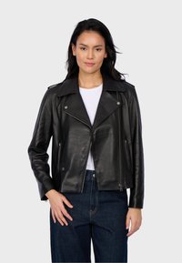 Veste de motard en cuir noir avec un large col, quincaillerie en argent, poches zippées et une silhouette ajustée, associée à une chemise blanche et un jean foncé.