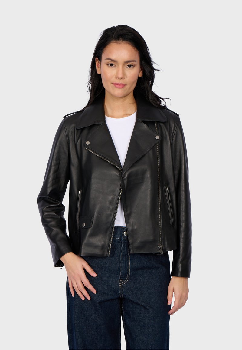 Veste de motard en cuir noir avec un large col, quincaillerie en argent, poches zippées et une silhouette ajustée, associée à une chemise blanche et un jean foncé.