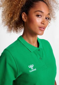 Grön polo skjorta i bomull, med krage, två knappar och en vit broderad logotyp på vänster bröst. Tyget har en strukturerad yta.