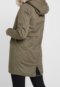 Olivgrüner Parka mit einer Kapuze aus Pelzbesatz, langen Ärmeln, tailliertem Schnitt und seitlichen Schlitzen am Saum; aus strapazierfähigem Stoff gefertigt.