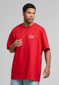 Rød bomulds t-shirt med løs pasform, prydet med hvid tekst "Dead WRONG" på venstre bryst. Enkel design med korte ærmer, ingen mønstre eller accenter.