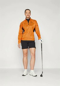 Orange lightweight jacket with black accents, zipper neck, paired with black shorts and white golf shoes, holding a golf club.