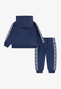 Ensemble de survêtement bleu marine comprenant une veste à capuche et un pantalon assorti. Les deux pièces comportent des accents à rayures blanches avec logo et des poignets côtelés.