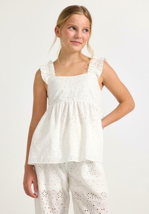 Pige iført hvid ærmeløs top med flæser og matchende bukser med hulmønster, stående med hånden på hoften foran en ensfarvet baggrund.