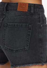 Pantalones cortos de mezclilla negra con dobladillo deshilachado, presentan una etiqueta de cuero marrón en la cintura y un único bolsillo trasero con detalles de costura visibles.