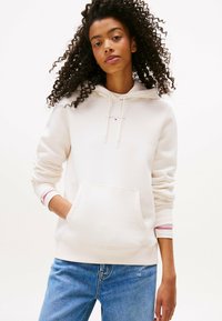 Giovane donna con capelli ricci che indossa un hoodie bianco sporco con tasca a marsupio e jeans blu, in piedi contro uno sfondo neutro.