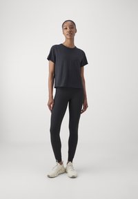 Czarny krótki top z krótkim rękawem, w połączeniu z czarnymi legginsami i jasnymi sneakerami. Materiał wydaje się gładki, z dopasowanym krojem. Styl minimalistyczny.