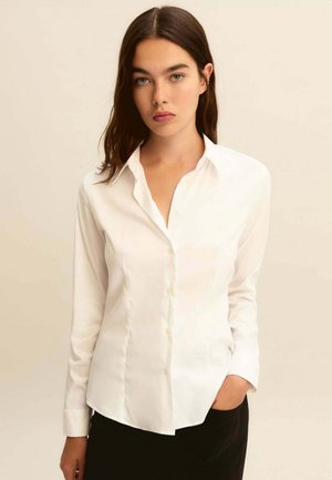 Giovane donna con lunghi capelli castani che indossa una camicia bianca aderente abbottonata e pantaloni neri, in piedi con una mano in tasca davanti a uno sfondo beige.