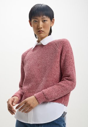 Maglione rosa a costine con maniche lunghe, scollo rotondo e tessuto testurizzato, indossato sopra una camicia bianca con colletto e jeans blu.