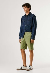 Άνδρας στέκεται φορώντας σκούρο μπλε πουκάμισο από τζιν με κουμπιά, χακί σορτς μέχρι το γόνατο και λευκά αθλητικά παπούτσια με σκουρόχρωμες λεπτομέρειες σε απλό φόντο.