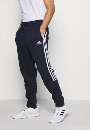 Granatowe dresy Adidas z białymi paskami po bokach, elastycznym pasem i kieszeniami. Noszone z białymi butami sportowymi z zielonymi akcentami.