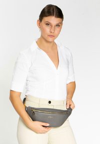 Grau Leder Gürteltasche mit einem goldenen Reißverschluss, mit geschwungener Form und strukturierter Oberfläche, gehalten von einer Person, die ein weißes Hemd und beigefarbene Hosen trägt.