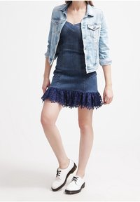 Niebieska sukienka dżinsowa z koronkowym wykończeniem, zestawiona z jasnoniebieską kurtką dżinsową i białymi butami. Posiada bezrękawnikowy krój i dopasowaną sylwetkę.