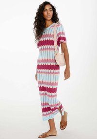 Sukienka maxi z dzianiny z zygzakowymi paskami w kolorze różowym, czerwonym i niebieskim. Sukienka ma krótkie rękawy, boczne rozcięcia i luźny krój. Noszona z płaskimi sandałami.