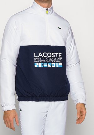 Mann trägt einen weißen und marineblauen Lacoste-Pullover mit Viertelreißverschluss, Koordinaten und Logos auf der Brust, kombiniert mit weißen Hosen.