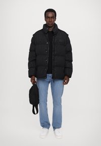 Black puffer jacket with a high collar, button closure, and textured surface. Worn over a black top with light blue jeans and white sneakers.