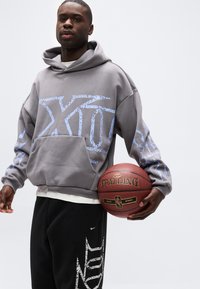 Νέος άνδρας που φοράει γκρι φούτερ με κουκούλα και μαύρο παντελόνι κρατάει μια μπάλα μπάσκετ Spalding μπροστά σε ένα απλό φωτεινό φόντο.