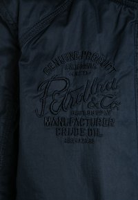 Dark blue fabric with black embroidered text reading "Genuine Product California - 8173 - Petrol Ind & Co. Blue Supply Manufacturer Crude Oil."