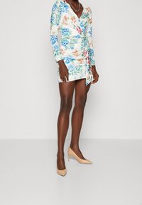 Robe portefeuille florale à manches longues, ornée de motifs bleus et roses sur un fond blanc. Portée avec des chaussures à talons beiges.