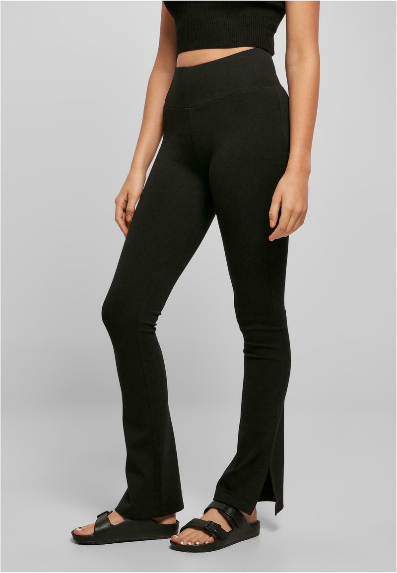 Femme portant des leggings noirs évasés à taille haute avec fentes latérales et sandales noires à boucle, debout sur un fond uni.