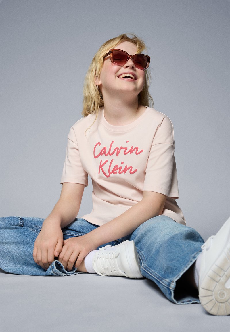 T-shirt rosa a maniche corte con il logo "Calvin Klein" rosso, abbinato a jeans in denim blu chiaro e sneaker bianche. Occhiali da sole marroni indossati.