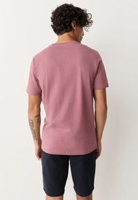 Kortærmet, rundhalset t-shirt i blød mauve bomuld, kombineret med mørkeblå shorts. Synlig tatovering på venstre underarm.