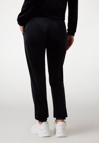 Zwarte fluwelen broek met een slanke pasvorm, elastische tailleband en enkellengte. Gecombineerd met witte sneakers voor contrast. Glad oppervlak, minimale details.