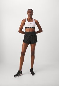 White sports bra with black logo band, black athletic shorts, and black sneakers with white accents. Model stands with hands on hips.
