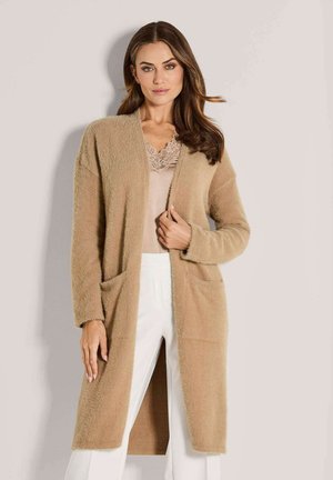 Vrouw in een beige lange fuzzy cardigan over een top met kantafwerking en witte broek, staande tegen een effen lichte achtergrond.