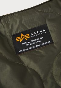 Tissu en nylon vert olive avec une texture matelassée. L'étiquette noire présente le logo "Alpha Industries" en orange, indiquant les détails du fabricant.