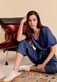 Denim jumpsuit met pofmouwen en een ceintuur om de taille, gecombineerd met crèmewitte sneakers met mesh en marineblauwe accenten. Op de achtergrond staat een vintage scooter.